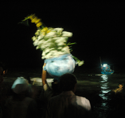 Imagem do ensaio Corpos e flores para Iemanjá, do fotógrafo Paulo Rossi, realizado na Praia de Tambaú, João Pessoa - PB. Na foto, os fieis são registrados, de costas, à madrugada, caminhando em diração ao mar, e levando sobre a cabeça um barco azul, enfeitado com flores, fitas e outros adereços, uma oferenda à Iemanjá, orixá das religiões de origem africana, rainha dos mares, padroeira dos pescadores e barqueiros.